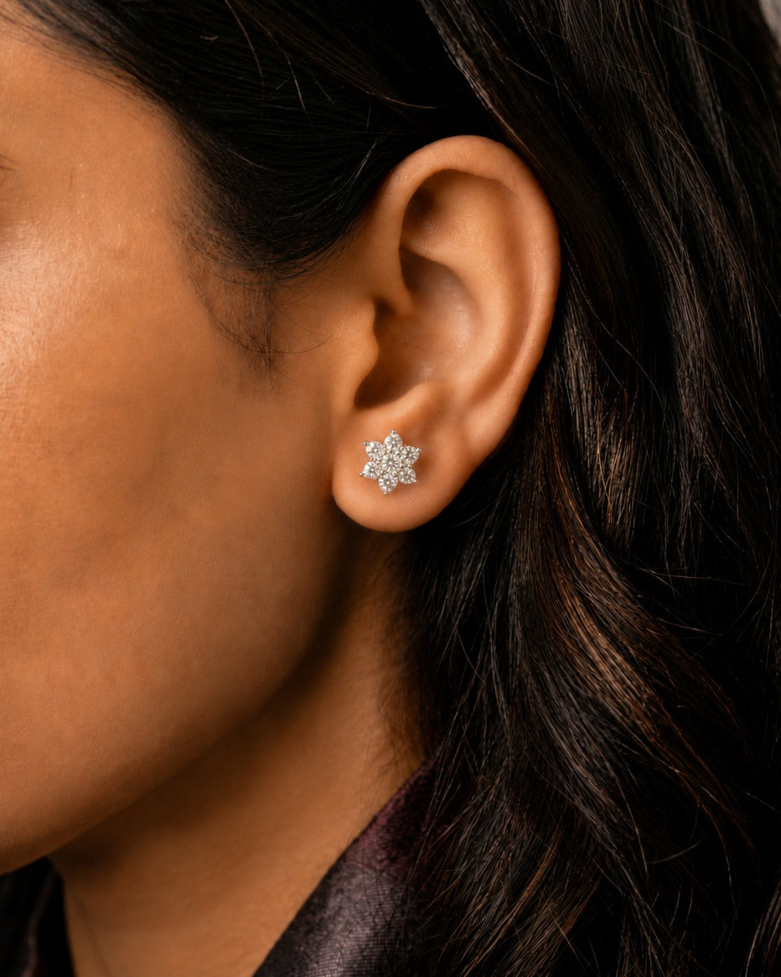 A close-up of a model's ear wearing a small silver-toned flower-shaped stud earring with clear stones.
