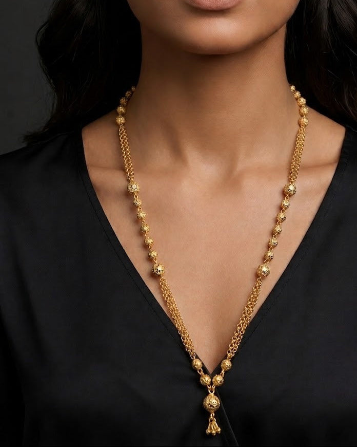 A gold long necklace with a beaded chain and a central pendant, shown on a model wearing a black satin V-neck top against a dark background.