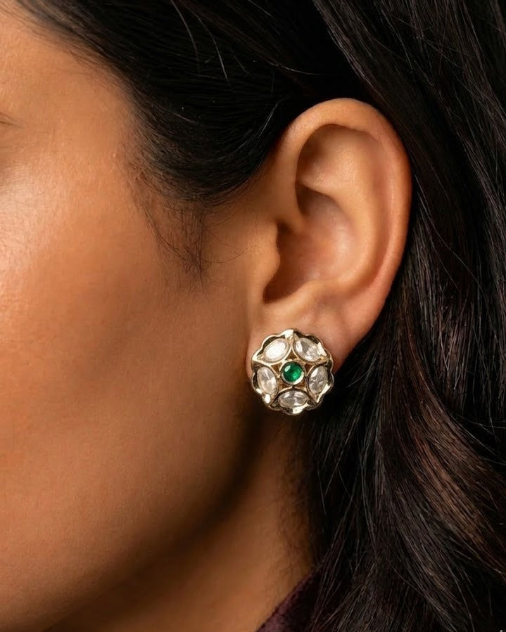 A close-up of a woman's ear wearing a detailed gold-toned floral stud earring with a central green stone and surrounding clear stones.