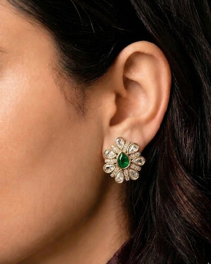 A close-up of a woman's ear wearing a gold flower-shaped stud earring with a green teardrop center and clear round stones.