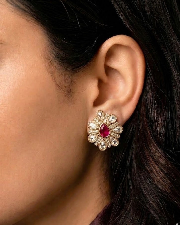 A close-up of a woman's ear wearing a gold-toned flower-shaped stud earring with a central red gemstone and surrounding clear stones.