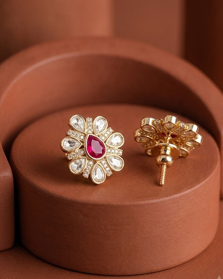 A pair of gold floral stud earrings with a central red gemstone and surrounding clear stones, displayed on a terracotta-colored circular pedestal.
