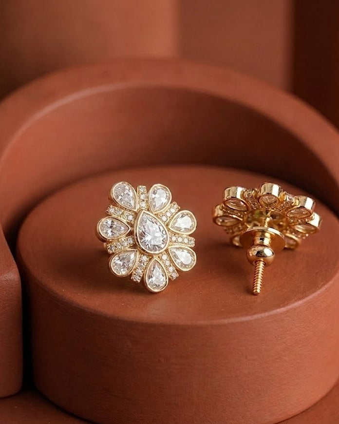 A pair of gold flower-shaped stud earrings with clear teardrop and round stones, displayed on a terracotta-colored circular pedestal.