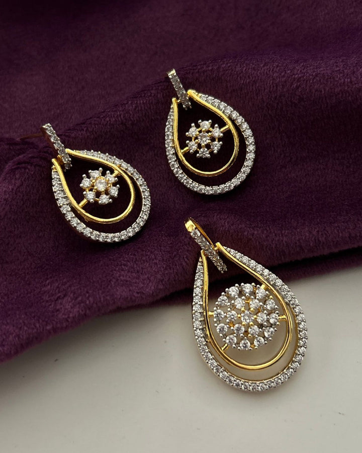 A pair of gold and silver teardrop-shaped earrings with pavé crystal borders and floral center details, displayed on a deep purple velvet fabric next to a light surface.