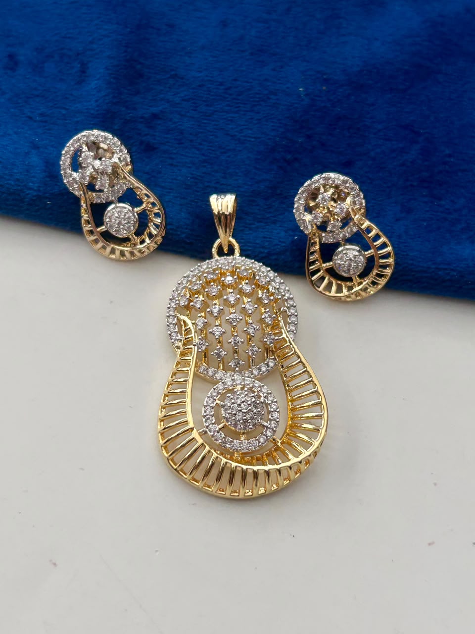 Gold and silver-tone jewelry set featuring a teardrop-shaped pendant and matching stud earrings, embellished with clear pavé stones, displayed on a white surface with a blue velvet background.