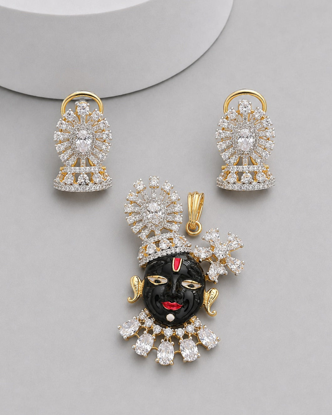 Gold-plated jewelry set featuring a black clay face pendant with red lips and forehead detail, surrounded by clear crystals, accompanied by matching crystal earrings on a light gray background.