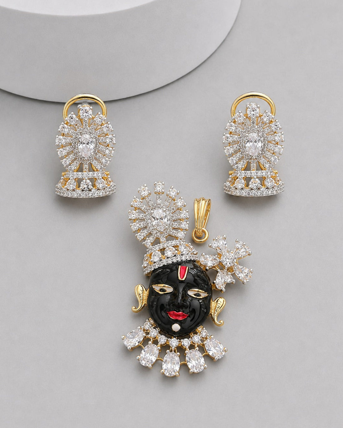 Gold-plated jewelry set featuring a black clay face pendant with red lips and forehead detail, surrounded by clear crystals, accompanied by matching crystal earrings on a light gray background.