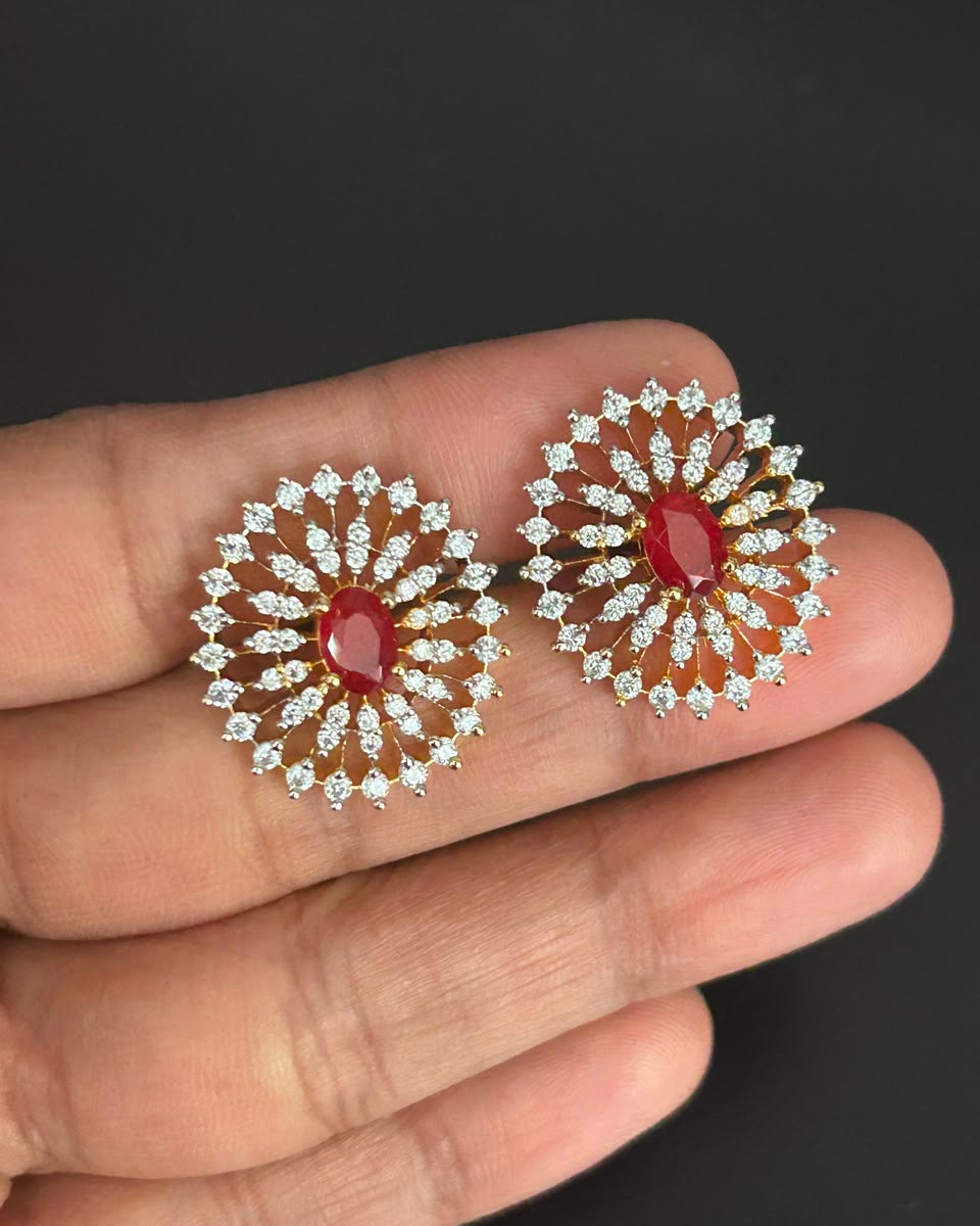 A pair of gold and silver floral stud earrings with red oval center stones and clear round-cut accents, held on a person's fingers against a dark background.