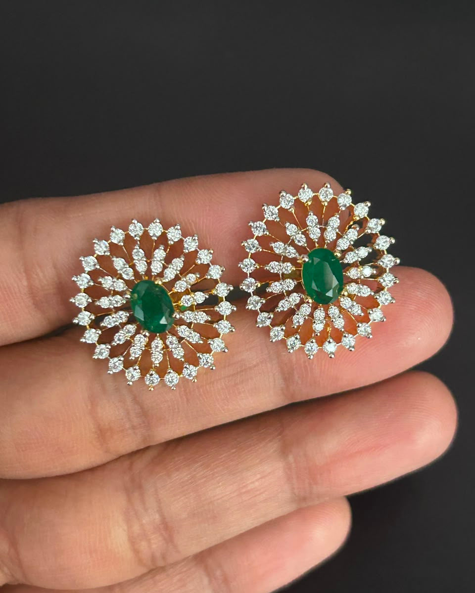 A pair of gold and diamond sunburst earrings with oval green emerald centers, held on a person's fingers against a dark background.