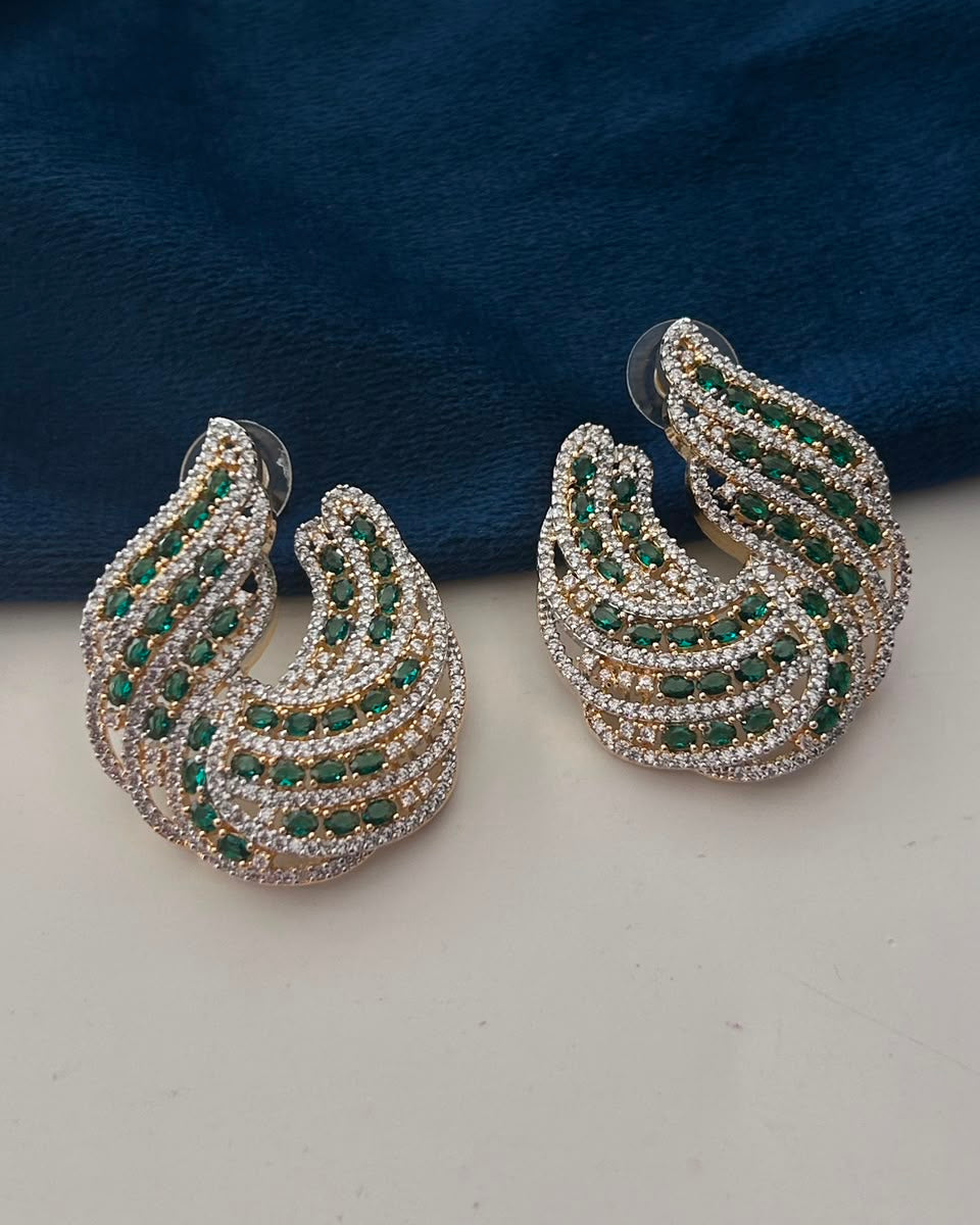 A pair of silver crescent-shaped hoop earrings embellished with rows of green and clear crystals, displayed on a light surface with a dark blue velvet background.