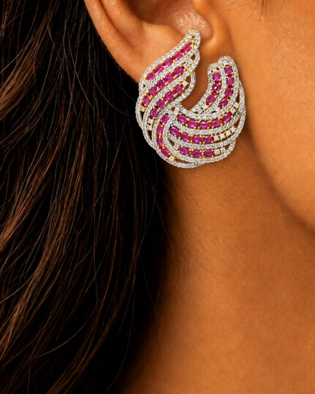 A close-up of a woman's ear wearing a statement wing-shaped stud earring set with bright pink gemstones and small clear stones on a silver-toned base.