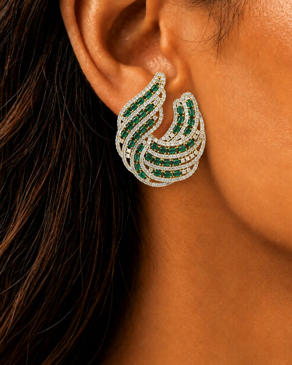 A close-up of a model's ear wearing a curved, multi-layered stud earring with green emeralds and clear pavé stones on a warm brown background.
