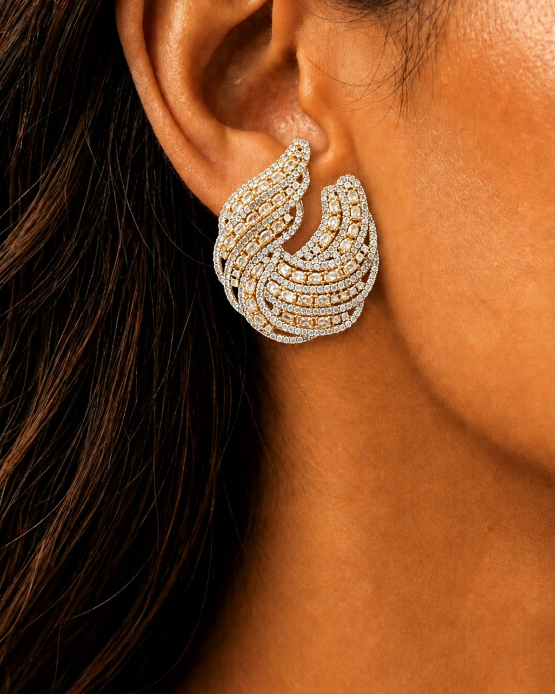 A close-up of a woman's ear wearing a large, multi-layered crescent-shaped earring set with small clear stones in a yellow-gold and silver-tone metal.