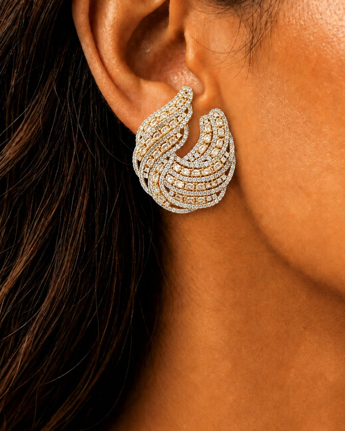 A close-up of a woman's ear wearing a large, multi-layered crescent-shaped earring set with small clear stones in a yellow-gold and silver-tone metal.