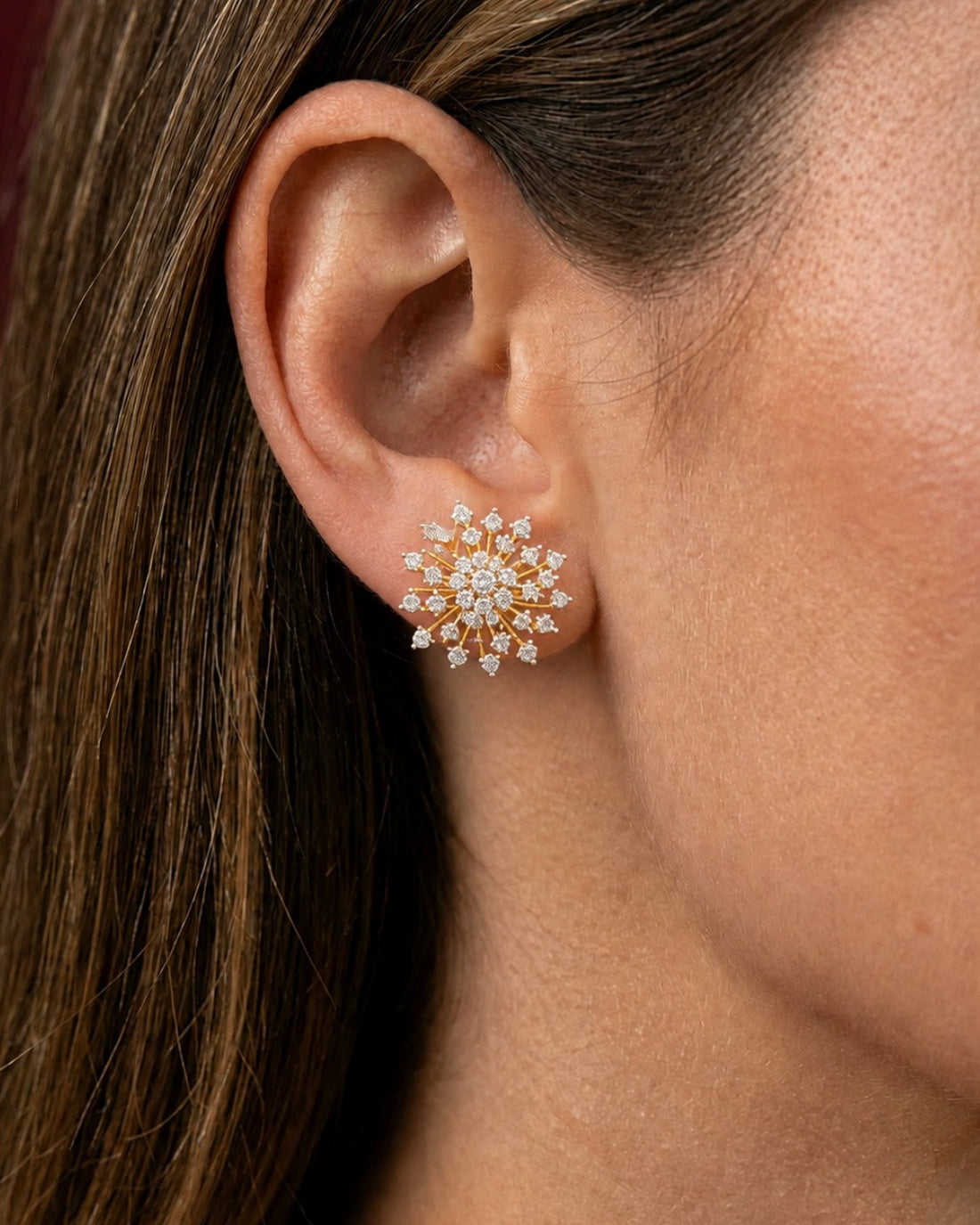 A close-up of a woman's ear wearing a delicate gold and diamond stud earring with a starburst cluster design.