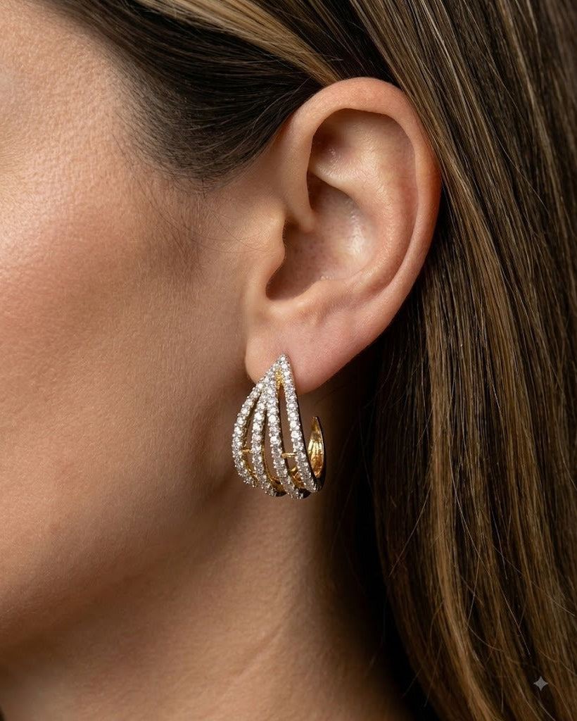A close-up of a model's ear wearing a gold huggie hoop earring adorned with multiple curved rows of sparkling clear pavé stones.