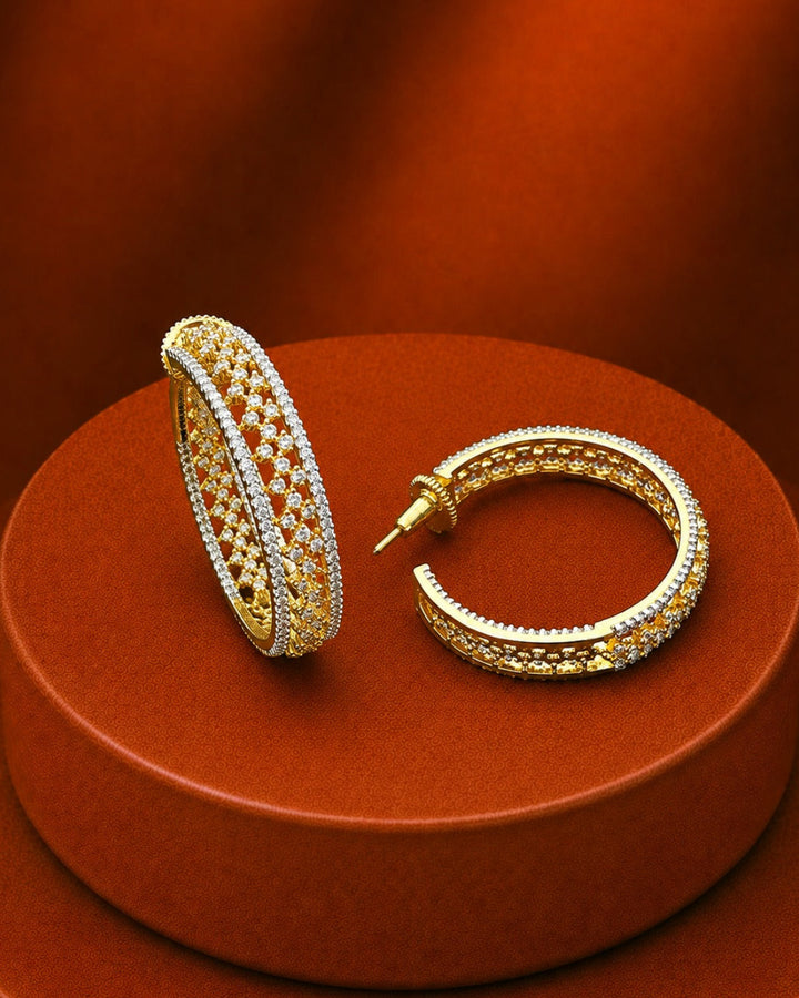 A pair of gold hoop earrings with intricate pavé-set clear stones, displayed on a round terracotta-colored pedestal against a matching warm brown background.