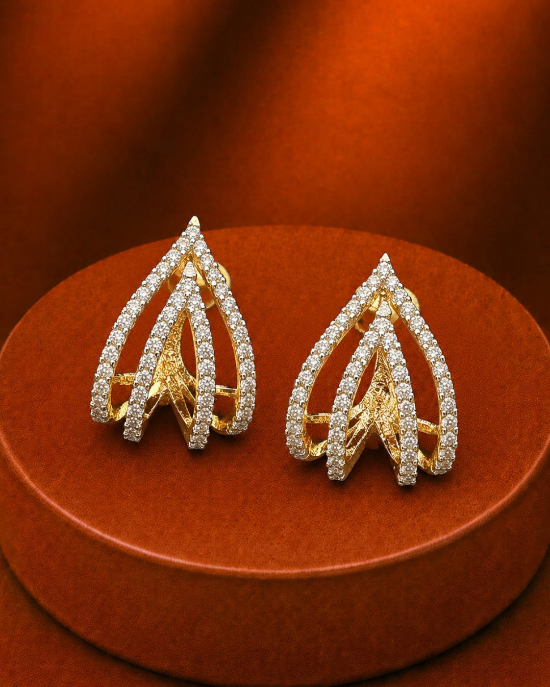 A pair of gold and diamond teardrop-shaped earrings displayed on a round brown jewelry stand against a warm brown background.