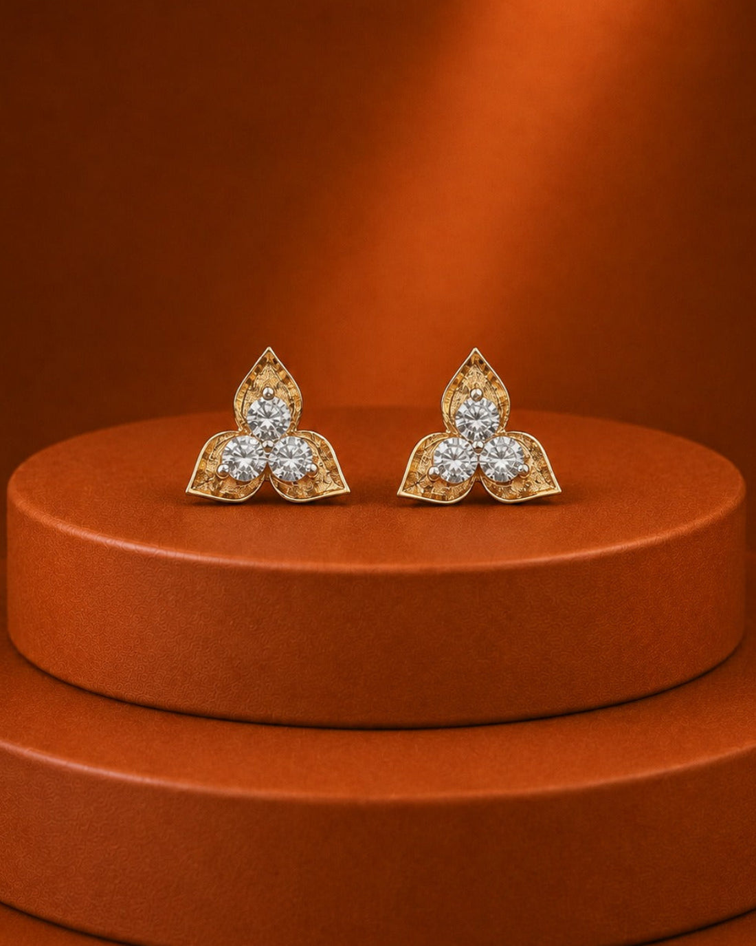 A pair of gold-toned butterfly stud earrings set with clear gemstones, displayed on a round terracotta pedestal against a matching warm brown background.