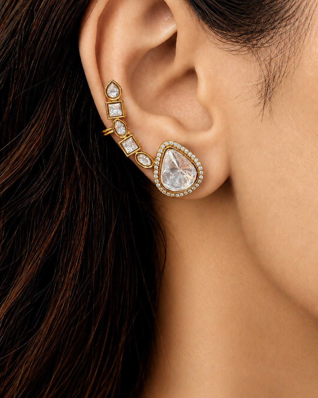 A close-up of a model's ear wearing a gold ear cuff with a large teardrop-shaped clear crystal and smaller clear crystals along with a row of small gold studs.