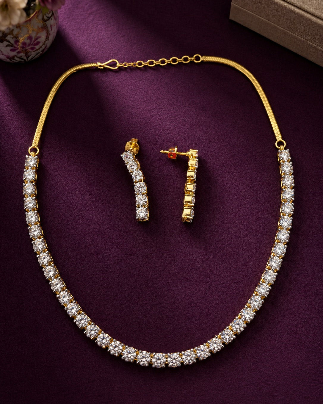 Gold necklace and matching earrings set with clear stones, arranged on a deep purple fabric background with a small decorative vase corner visible.