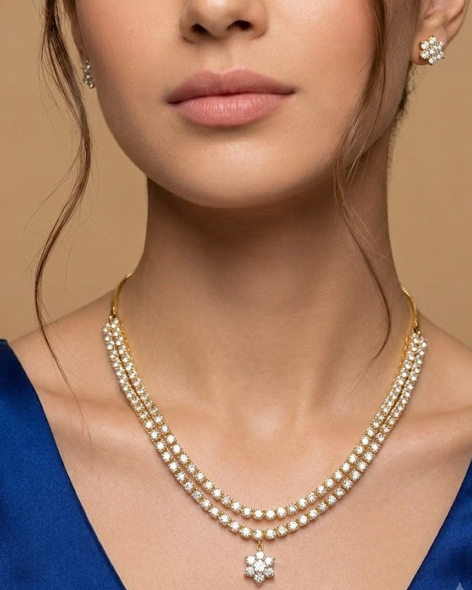 A close-up of a model wearing a double-row pearl necklace with a gold-tone flower-shaped pendant, matching flower stud earrings, and a blue satin top against a warm beige background.