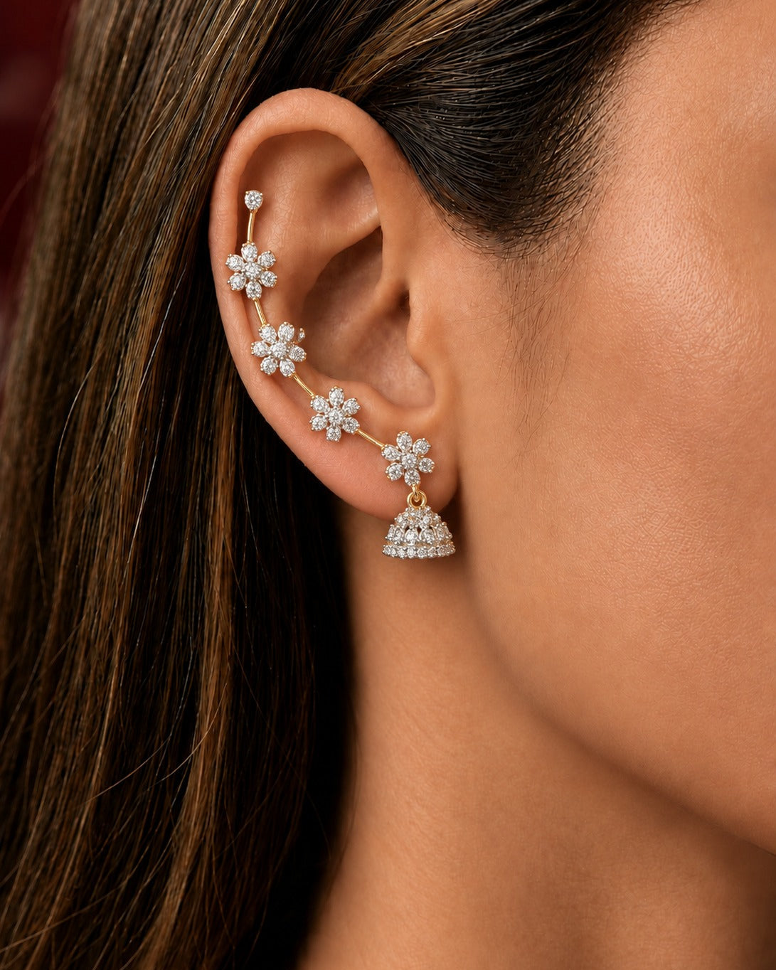 A close-up of a woman's ear wearing a gold ear cuff adorned with four silver flower-shaped studs and a dangling silver bell-shaped charm set with clear stones.