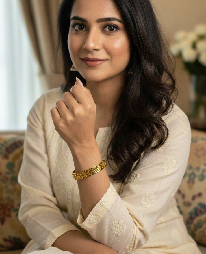 Woman in a white traditional outfit with gold jewelry, sitting indoors.