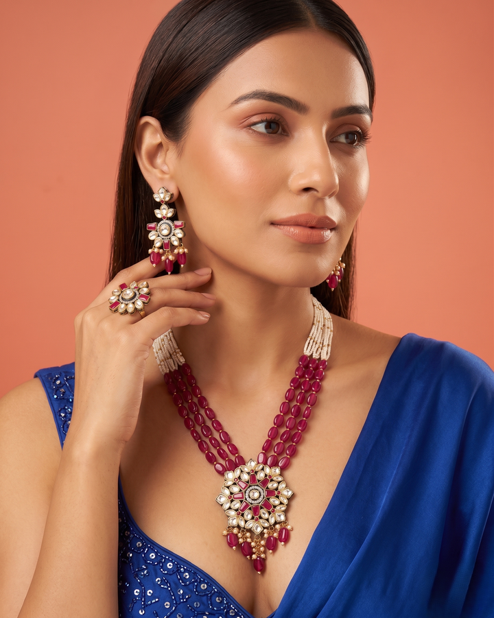 Woman wearing a blue saree with a red and gold necklace and earrings against a peach background