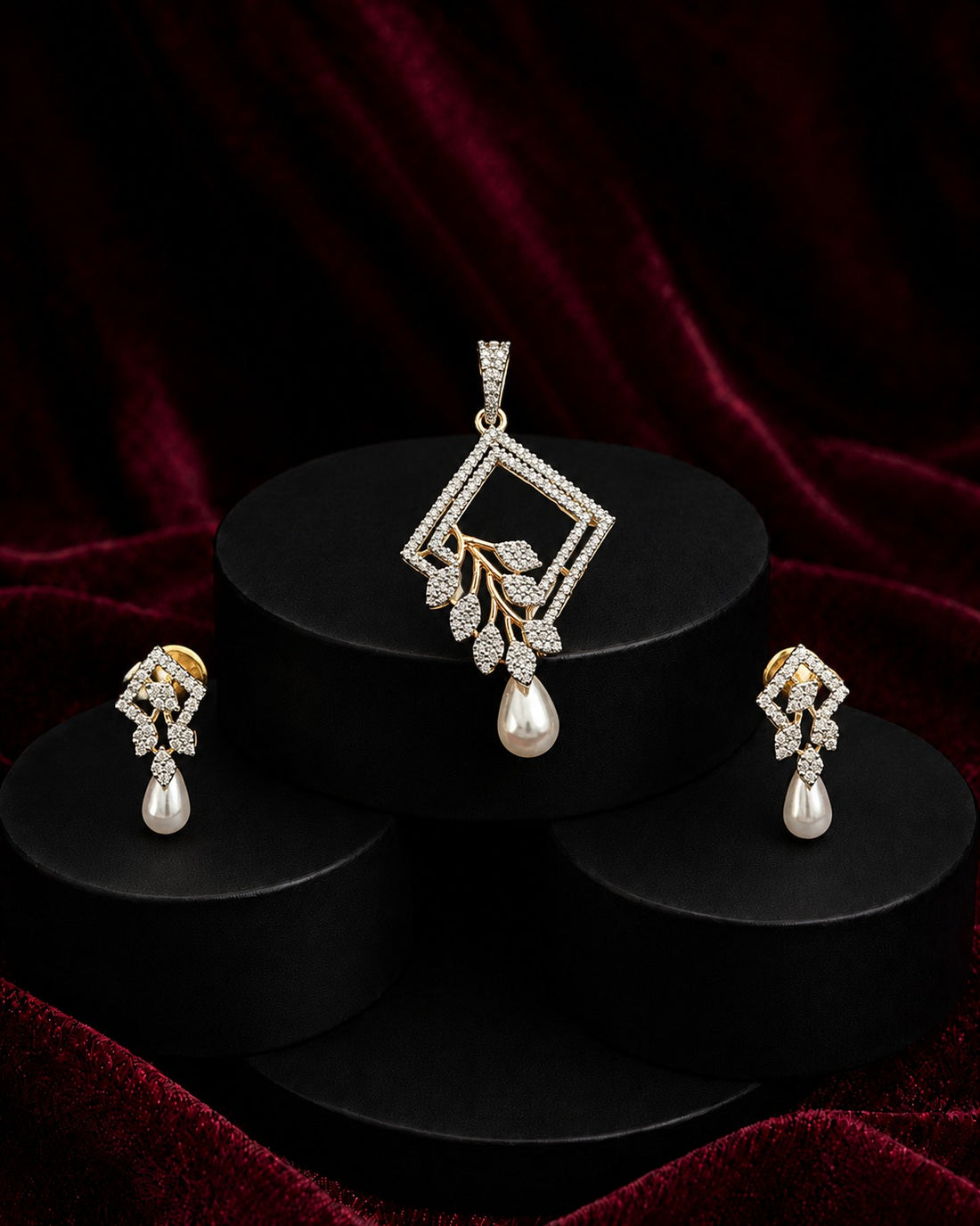A jewelry set featuring a diamond-shaped pendant with a dangling pearl and leaf motif, paired with matching drop earrings, displayed on black cylindrical pedestals against a dark red velvet background.