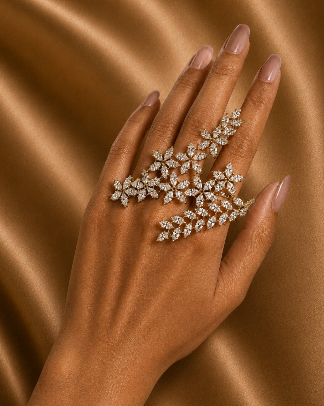 A close-up of a hand with manicured nails wearing a large silver-toned ring set with multiple clear stones in a leafy, floral cluster design, posed against a draped brown satin fabric background.
