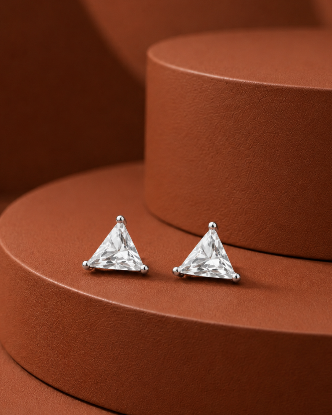 A pair of silver stud earrings with triangular clear gemstones, displayed on a textured terracotta-colored circular pedestal.
