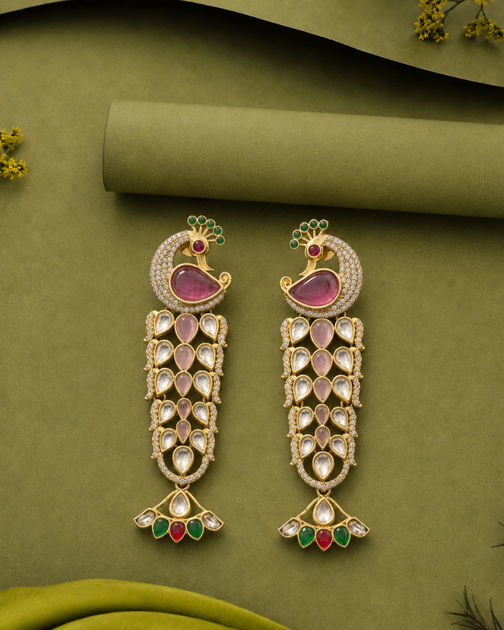 A pair of ornate gold-toned dangle earrings with pink stone inlays, clear rhinestone embellishments, and small green and red accents, arranged on an olive green fabric background with decorative paper and flowers.