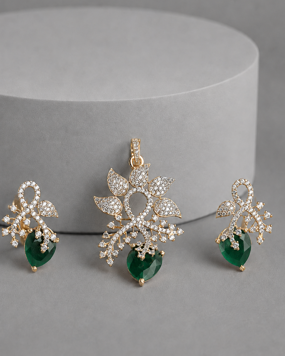 A gold-toned jewelry set featuring a central pendant and two matching drop earrings, decorated with pavé-set clear stones and a heart-shaped green gemstone at the bottom, displayed on a gray surface in front of a cylindrical pedestal.