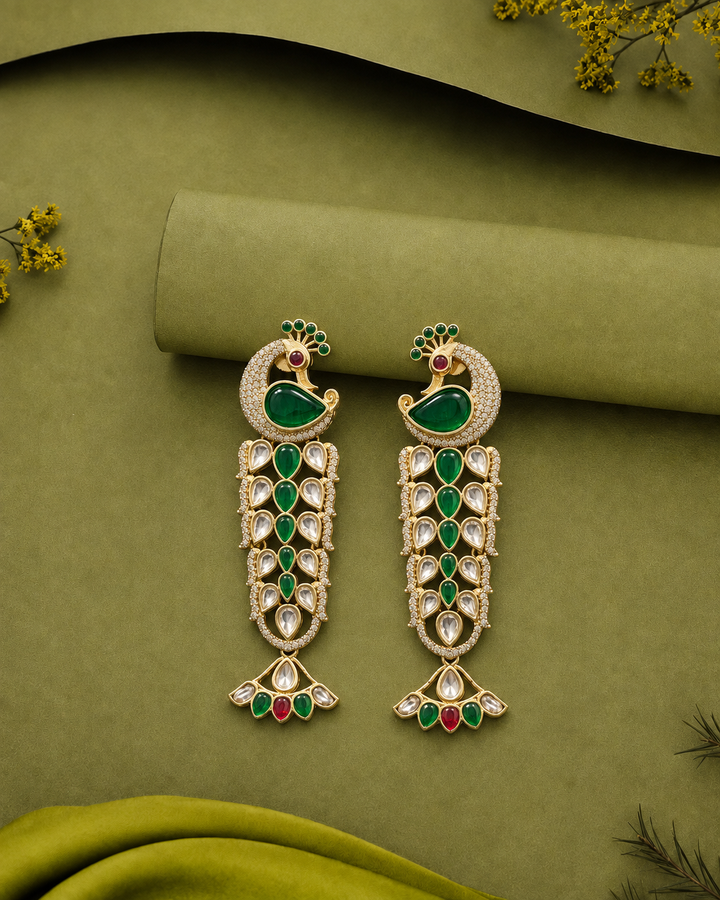 A pair of gold-plated peacock dangle earrings with green enamel, clear stones, and red accents, displayed on an olive green fabric background.
