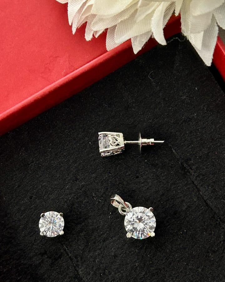A pair of silver stud earrings and a matching pendant with clear round-cut gemstones, displayed on a black velvet surface next to a red box and white fabric flower.