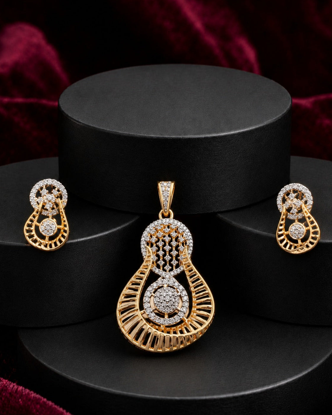 Gold jewelry set featuring a teardrop pendant, matching earrings, and a small box, all adorned with clear stones, displayed on a black tiered stand with a dark red velvet background.