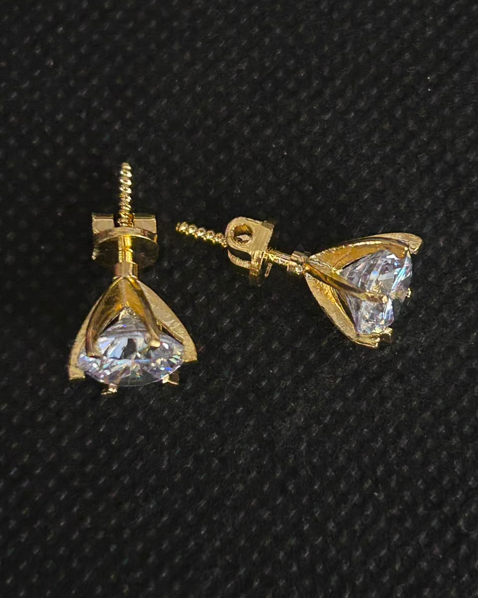 A pair of gold triangular stud earrings with clear faceted gemstones, displayed on a textured black background.