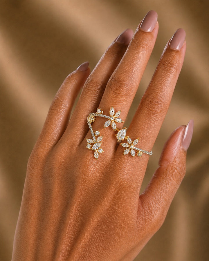 A close-up of a hand with manicured nails wearing a delicate gold ring set with small clear stones in a floral and leaf design, against a soft warm brown background.