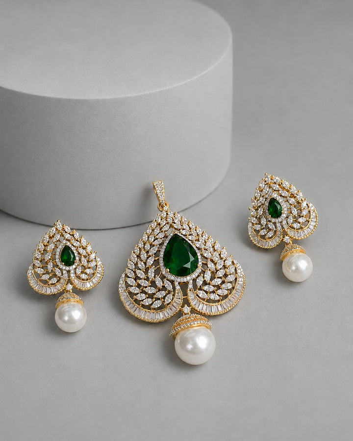 Gold-plated teardrop-shaped jewelry set with green teardrop gemstones, pavé-set clear crystals, and dangling white pearl beads, arranged on a gray surface with a white cylindrical prop.