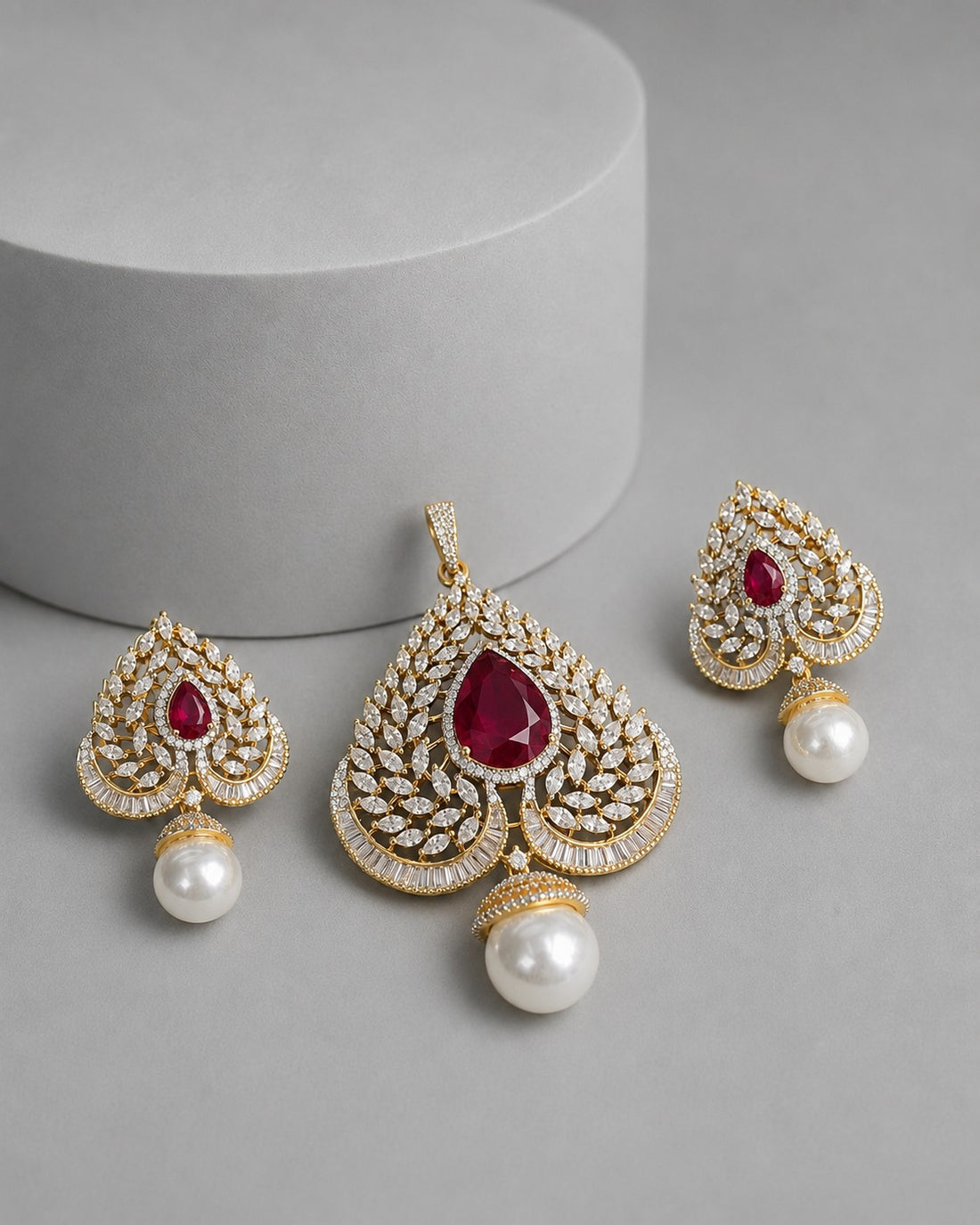 Gold-plated Indian style jewelry set featuring a large pendant and matching earrings, embellished with pavé-set clear stones and a central teardrop-shaped red gemstone, with a dangling white pearl at the bottom, displayed on a gray surface with a cylindrical prop.
