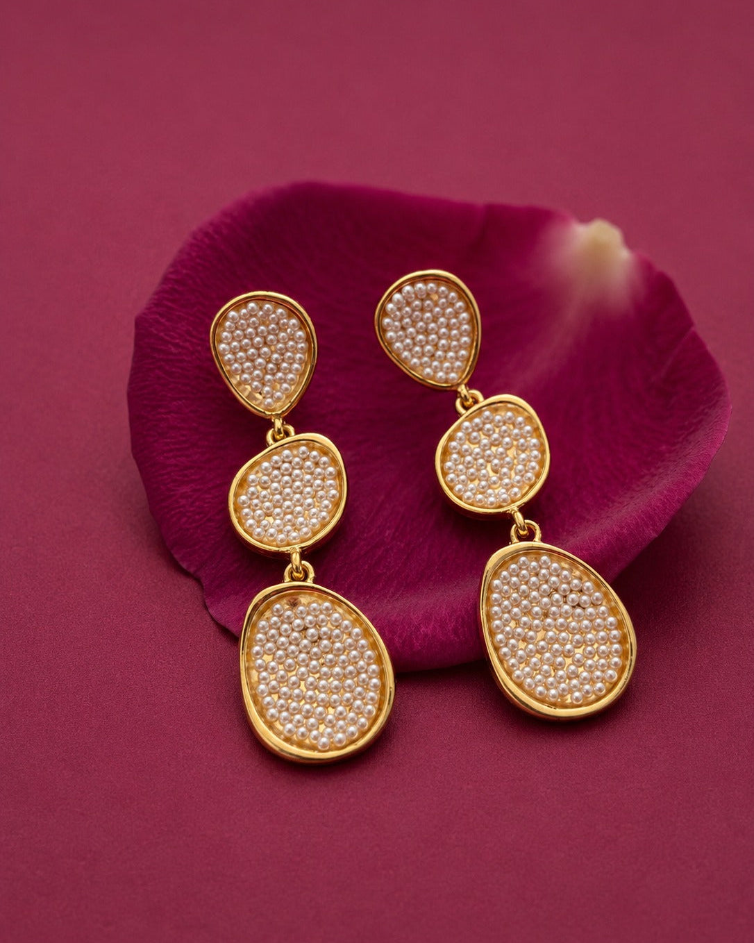A pair of gold drop earrings with teardrop-shaped settings filled with small white stones, displayed on a deep magenta flower petal against a matching magenta background.