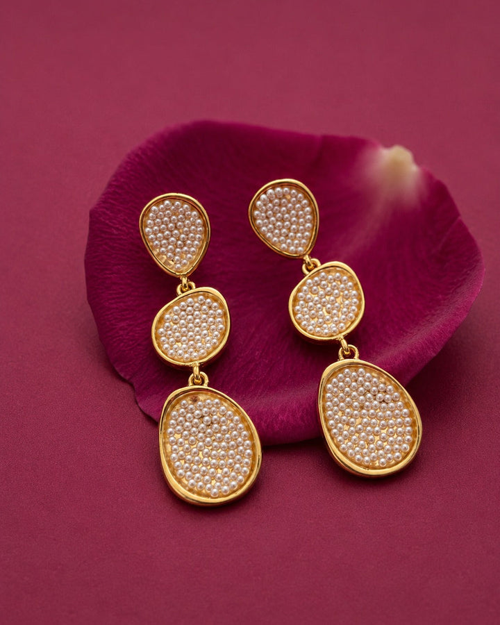 A pair of gold drop earrings with teardrop-shaped settings filled with small white stones, displayed on a deep magenta flower petal against a matching magenta background.