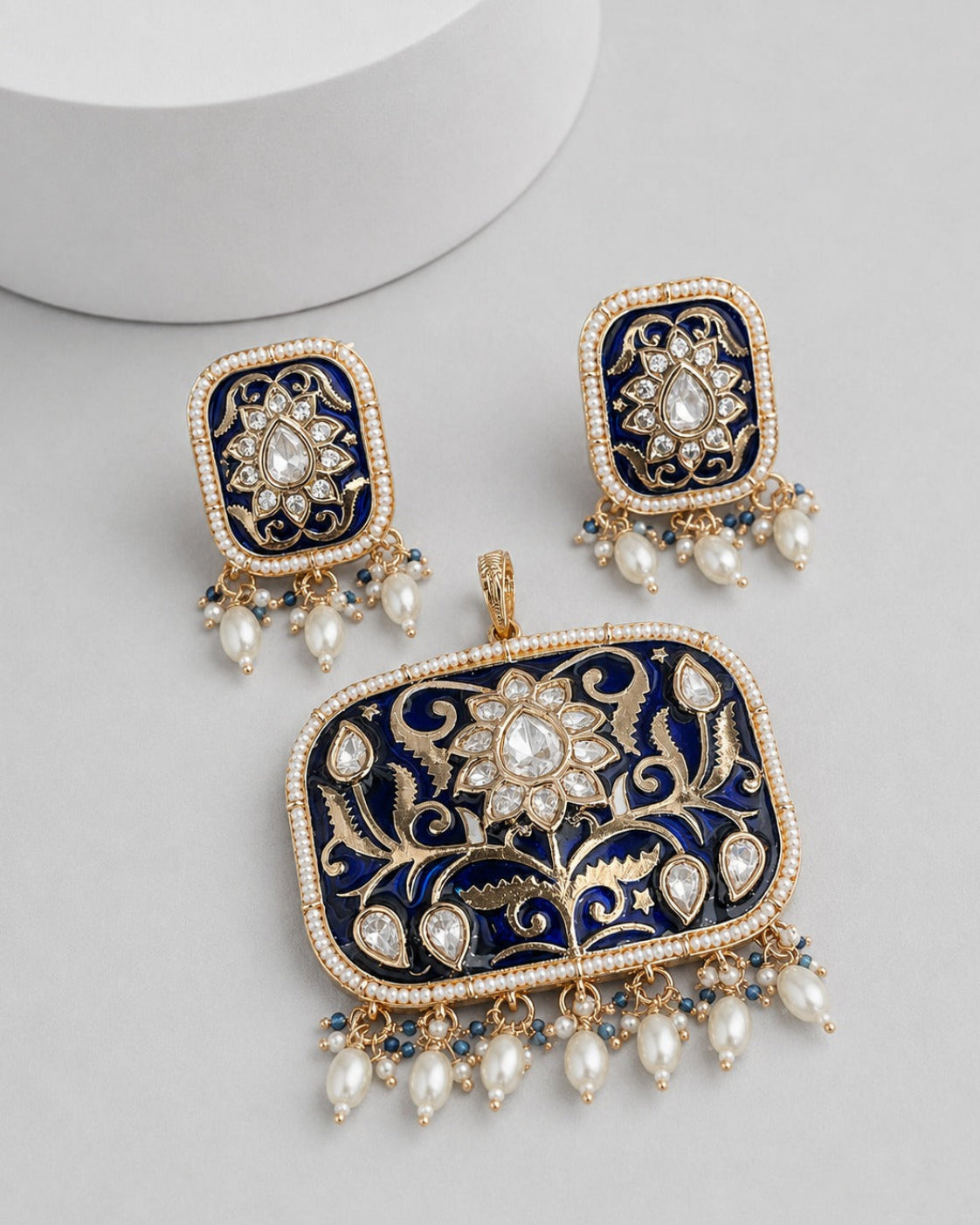 A pair of rectangular gold-toned statement earrings with a matching pendant, featuring deep navy blue enamel, intricate gold floral filigree, and dangling white faux pearls on a light gray background.