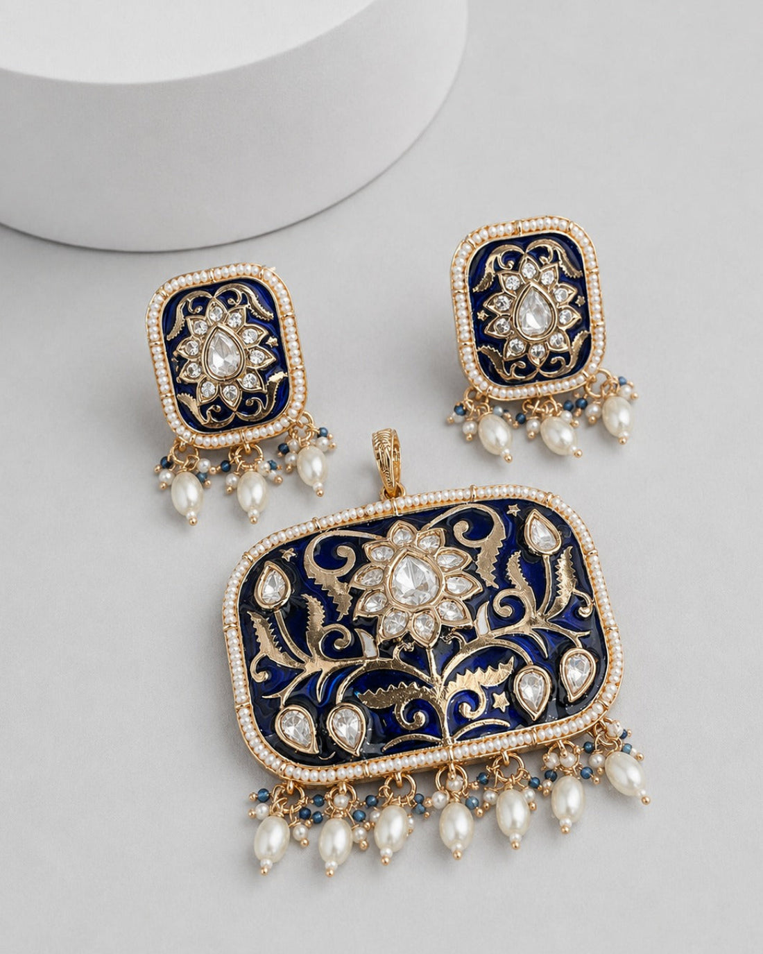 A pair of rectangular gold-toned statement earrings with a matching pendant, featuring deep navy blue enamel, intricate gold floral filigree, and dangling white faux pearls on a light gray background.