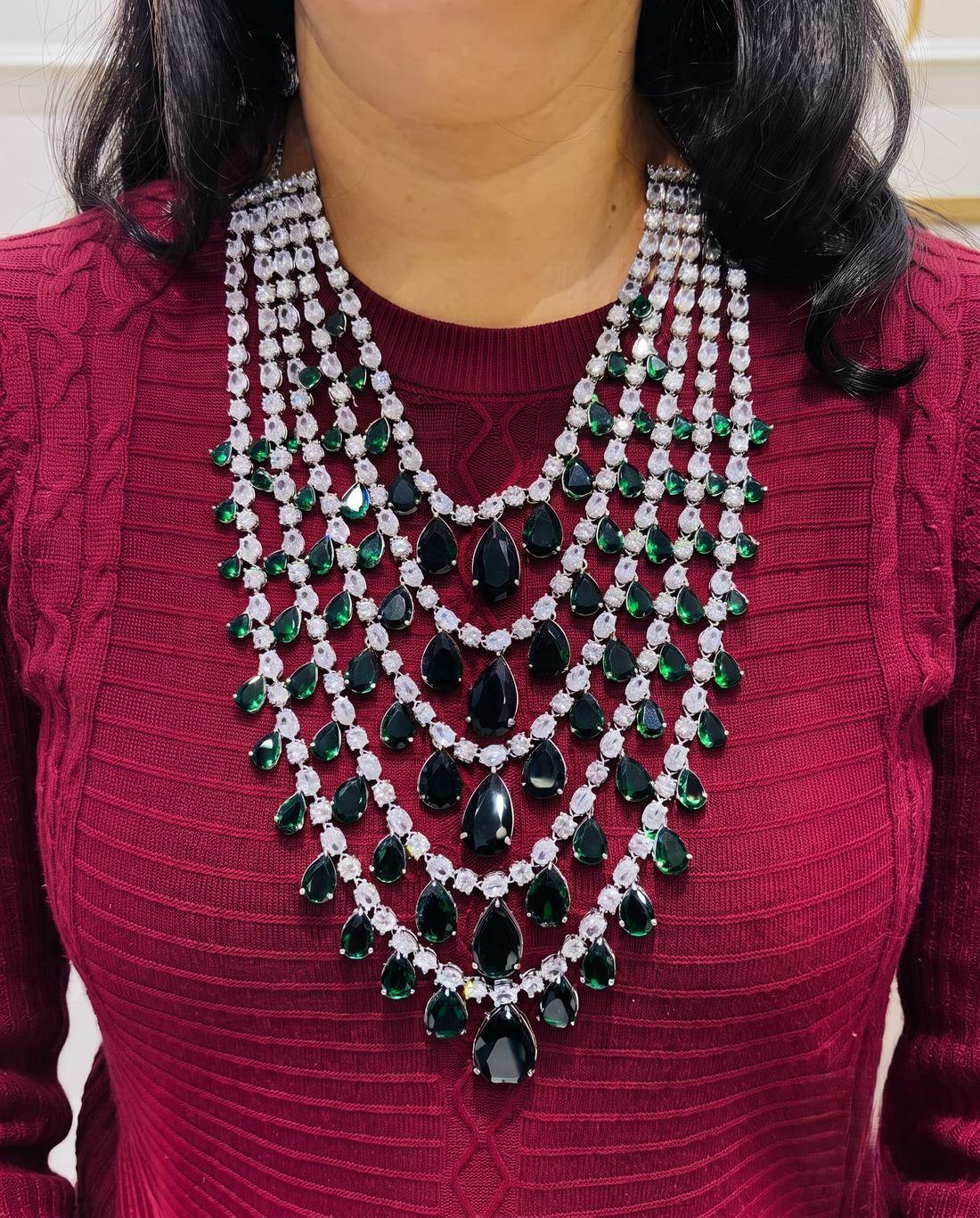 A close-up of a woman wearing a multi-strand statement necklace. The necklace features multiple layers of clear rhinestones and dark green teardrop and oval gemstones.