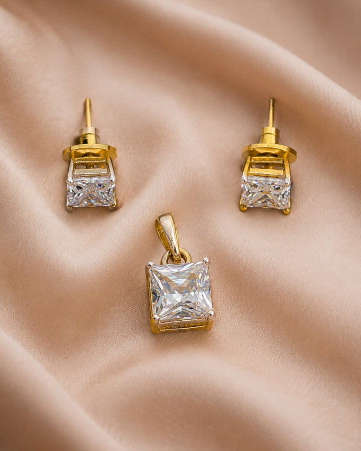 A pair of gold stud earrings and a matching pendant featuring clear princess-cut gemstones, arranged on a draped beige satin fabric background.