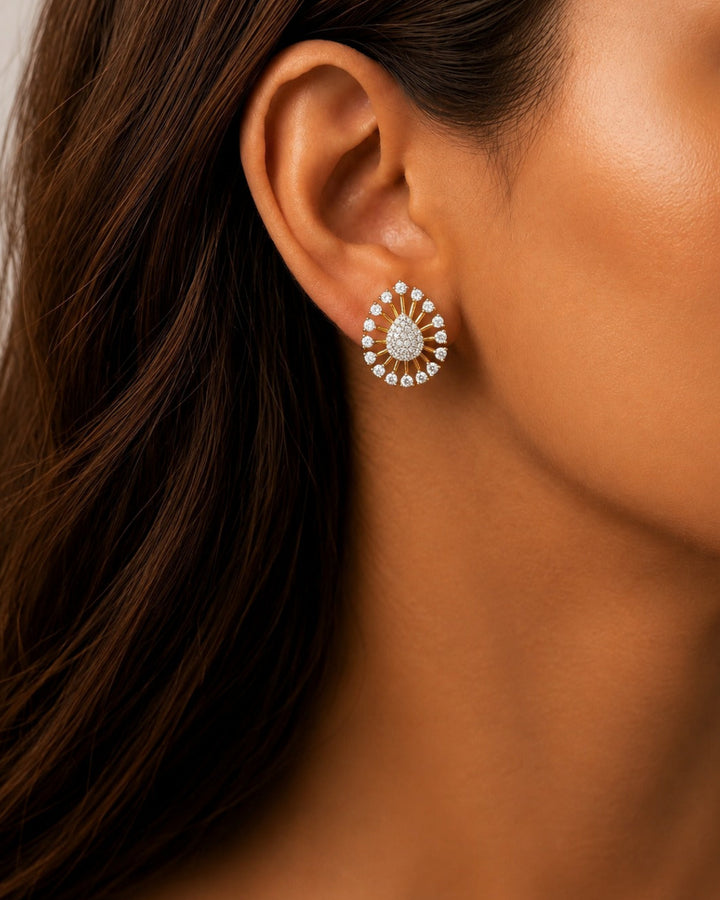 A close-up of a model's ear wearing a circular stud earring with a sunburst design made of clear stones in a yellow-gold setting.