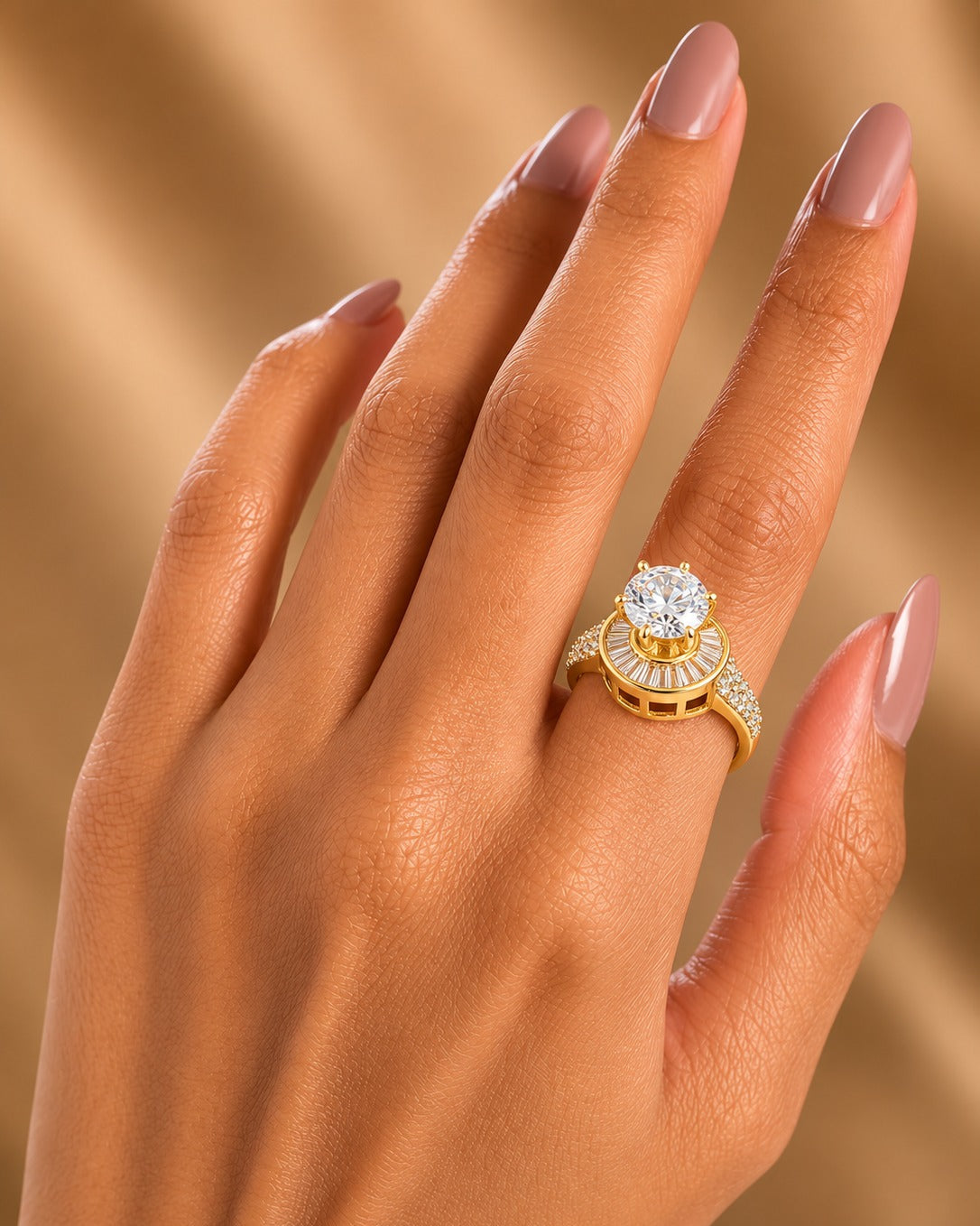 A close-up of a hand with nude manicured nails wearing a yellow gold ring featuring a large round-cut diamond and a halo of smaller diamonds.