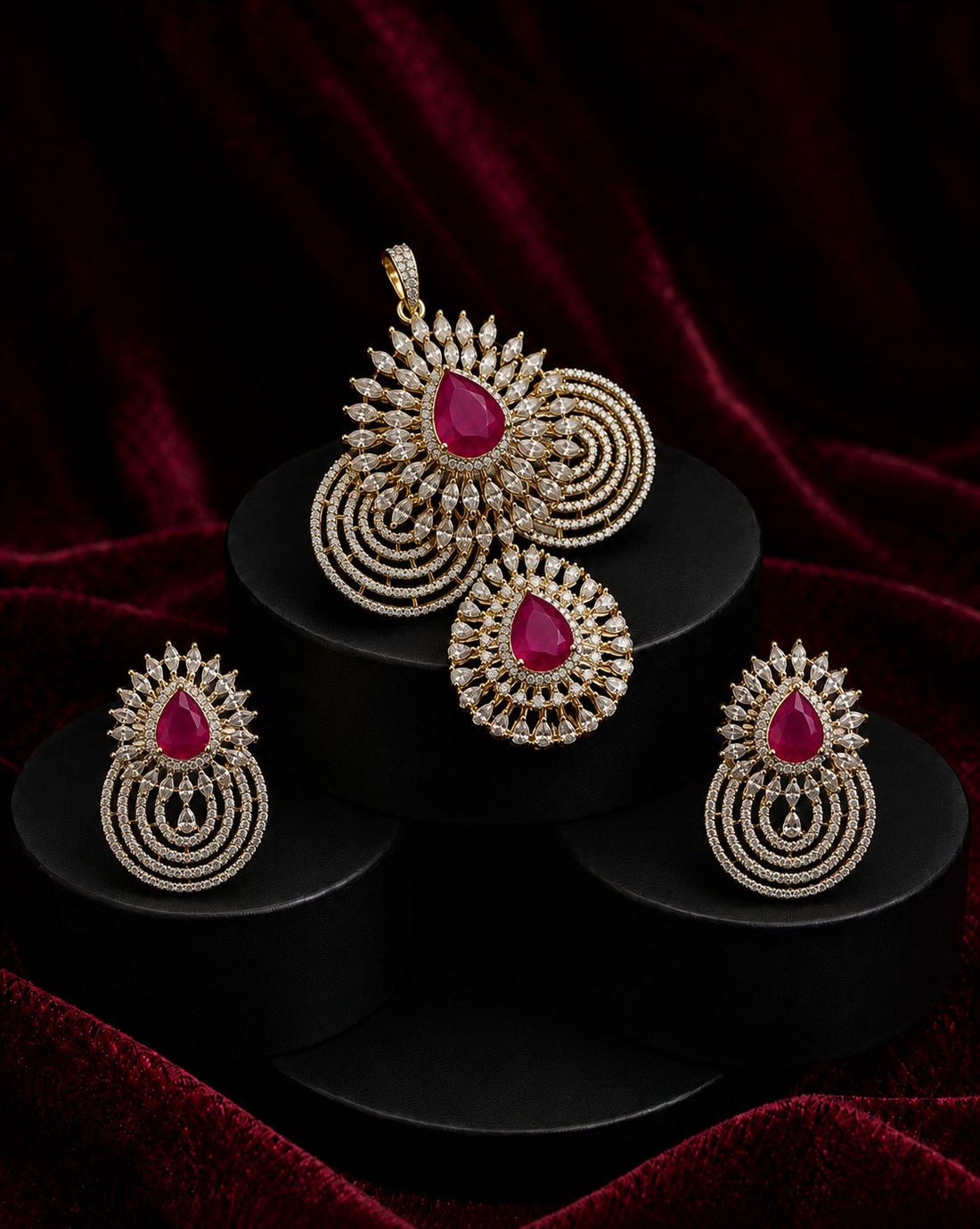 Gold-toned traditional Indian jewelry set featuring a pendant, earrings, and a large brooch, each adorned with a central teardrop-shaped pink gemstone and intricate circular gold pavé detailing, displayed on black pedestals against a dark red velvet background.